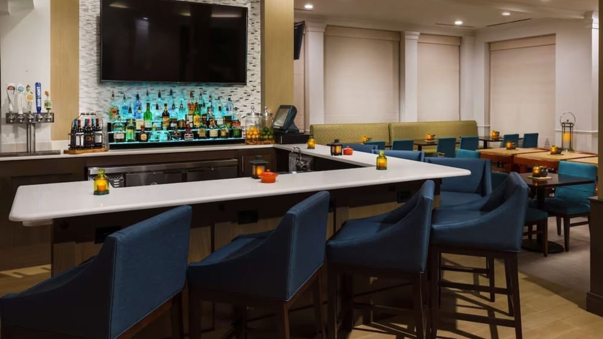 Stylish hotel bar with a white countertop, blue upholstered bar stools, illuminated liquor display, beer taps, large TV and adjacent dining tables.