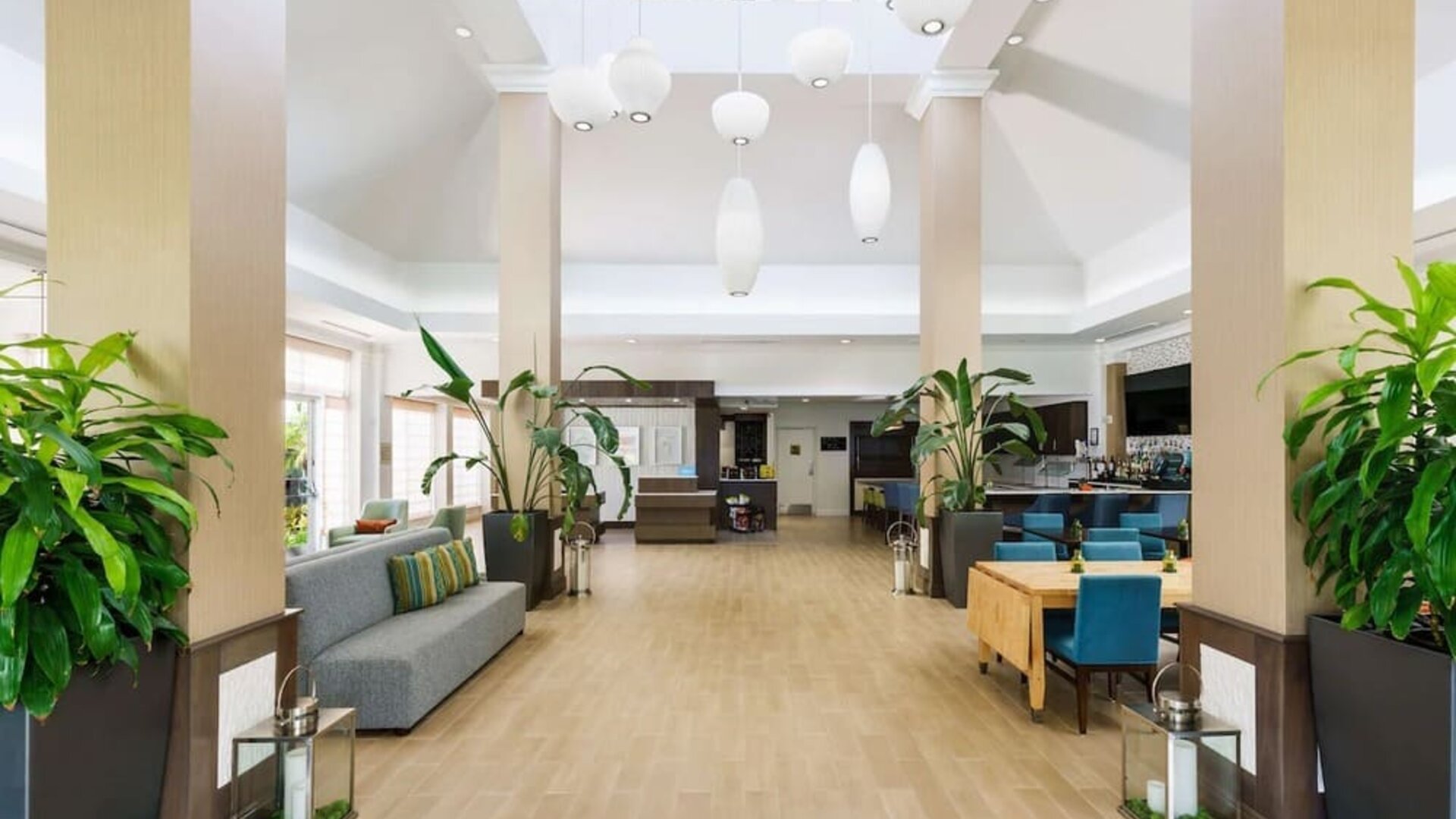 Bright, open hotel lobby with tall columns, large tropical plants, gray sofa seating, turquoise dining chairs, pendant lights and access to the bar and reception areas.