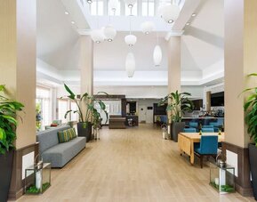 Bright, open hotel lobby with tall columns, large tropical plants, gray sofa seating, turquoise dining chairs, pendant lights and access to the bar and reception areas.