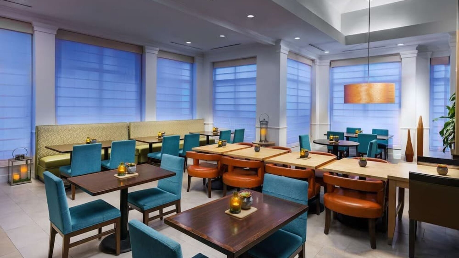 Contemporary hotel dining area with turquoise chairs, cushioned booth seating, dark wood tables, warm pendant lighting and large windows with privacy shades.