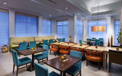 Contemporary hotel dining area with turquoise chairs, cushioned booth seating, dark wood tables, warm pendant lighting and large windows with privacy shades.