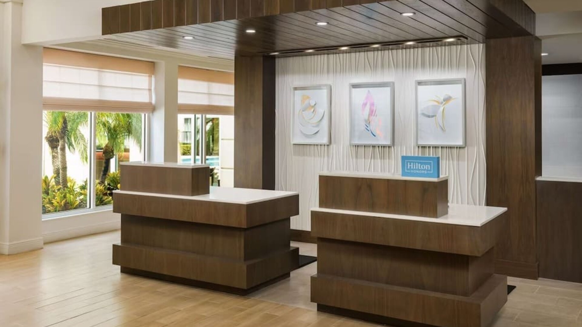 Modern hotel reception area with two wood-and-white front desks, contemporary artwork, Hilton Honors signage and large windows overlooking tropical landscaping.