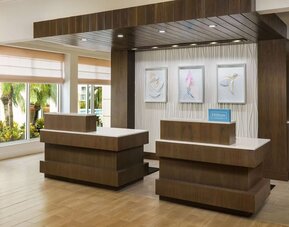 Modern hotel reception area with two wood-and-white front desks, contemporary artwork, Hilton Honors signage and large windows overlooking tropical landscaping.