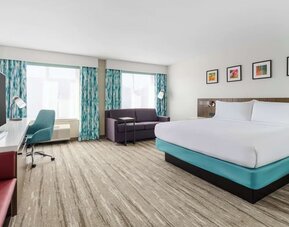 Spacious modern hotel room with a king bed, sofa, large TV, work desk, teal office chair, two large windows and colorful blue-patterned curtains.