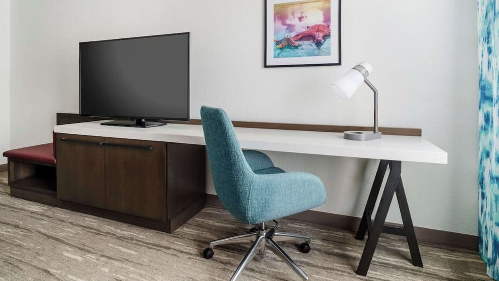 Hotel guest room workspace with a long white desk, teal rolling chair, desk lamp, large flat-screen TV, dark wood cabinet and colorful framed artwork.