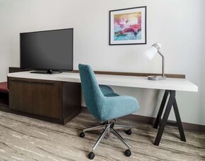 Hotel guest room workspace with a long white desk, teal rolling chair, desk lamp, large flat-screen TV, dark wood cabinet and colorful framed artwork.