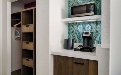 In-room coffee station with a single-serve coffee maker, microwave, ice bucket, storage cabinet, open closet, hangers and ironing board against a decorative blue accent wall.