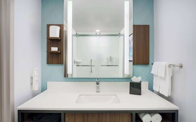 Modern hotel bathroom vanity with a white countertop and sink, large illuminated mirror, open towel storage and glass-enclosed shower reflected in the mirror.