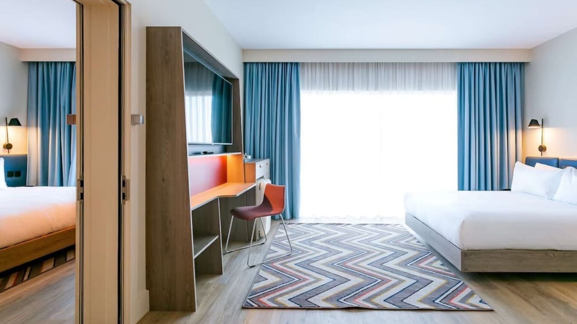 Spacious modern hotel guest room with king-size bed, floor-to-ceiling window with blue curtains, wall-mounted television, work desk and chair, wood flooring, and colorful geometric rug.