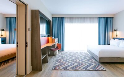 Spacious modern hotel guest room with king-size bed, floor-to-ceiling window with blue curtains, wall-mounted television, work desk and chair, wood flooring, and colorful geometric rug.