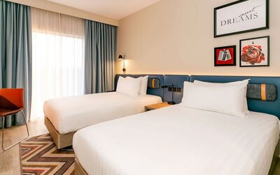 Bright contemporary hotel room with two beds, crisp white linens, blue upholstered headboard, framed wall art, floor-to-ceiling window with blue curtains, orange chair, wood flooring, and patterned rug.