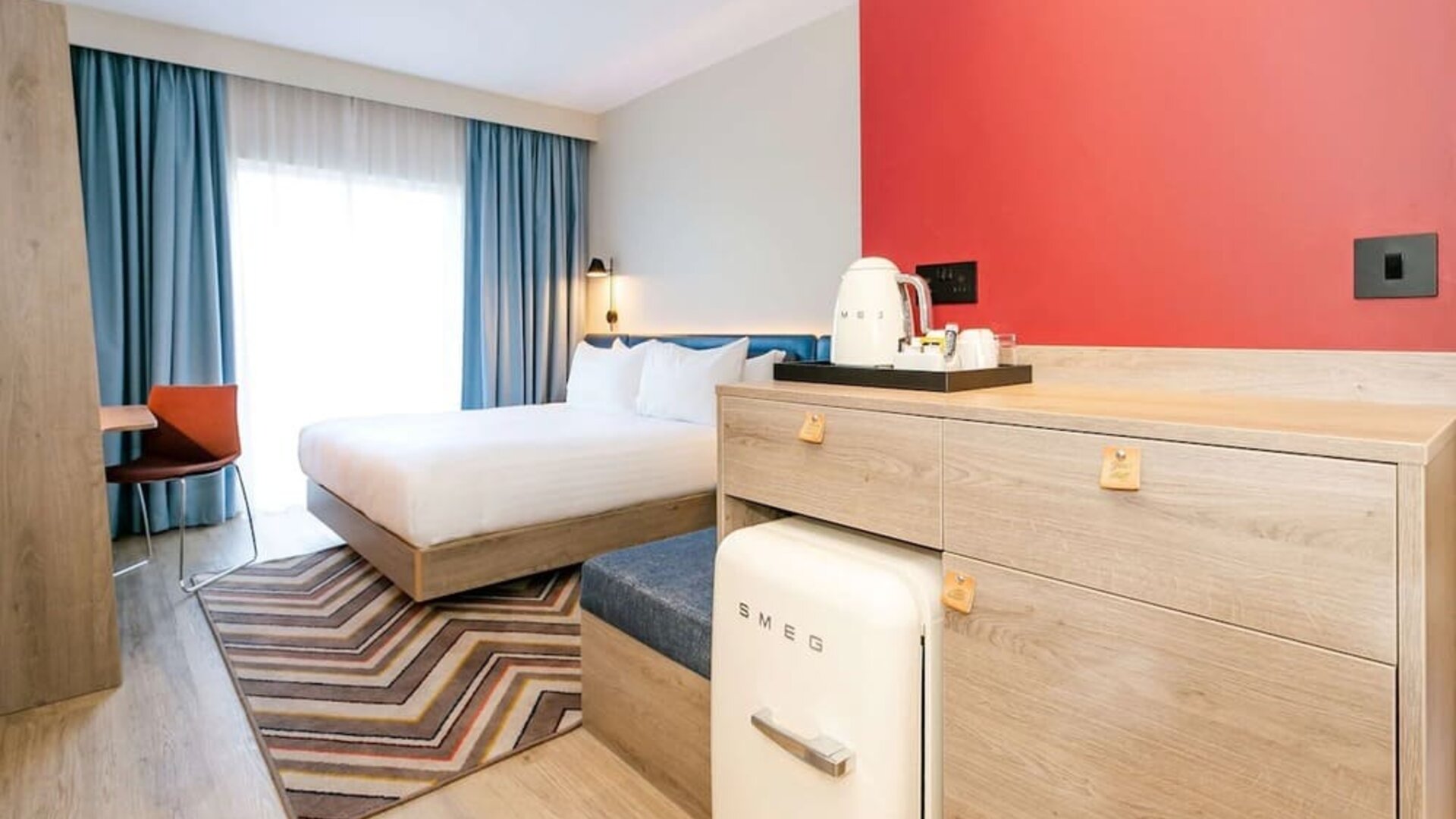 Modern hotel guest room with a king-size bed, white bedding, blue curtains, wood furnishings, red accent wall, compact SMEG refrigerator, tea and coffee station, patterned rug, and desk chair.
