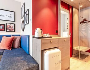 Modern hotel room amenity area with a blue upholstered bench, decorative cushions, framed artwork, tea and coffee station, compact SMEG refrigerator, open wardrobe with hangers, luggage rack, and red accent walls.