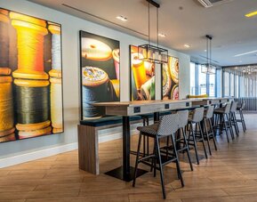 Contemporary hotel coworking space with a long communal wooden table, power outlets, patterned high stools, modern pendant lighting, and colorful wall artwork featuring spools of thread and textile imagery.