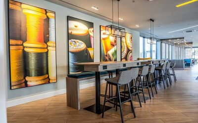 Contemporary hotel coworking space with a long communal wooden table, power outlets, patterned high stools, modern pendant lighting, and colorful wall artwork featuring spools of thread and textile imagery.