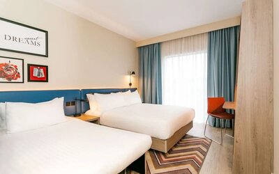 Bright modern hotel guest room with two beds dressed in white linens, a blue upholstered headboard, bedside lighting, floor-to-ceiling windows with blue curtains, a patterned rug, and a compact work desk.