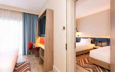 View between adjoining hotel sleeping areas featuring multiple beds with white linens, blue upholstered headboards, floor-to-ceiling windows with blue curtains, built-in desk and television unit, orange chair, wood flooring, and geometric rugs.