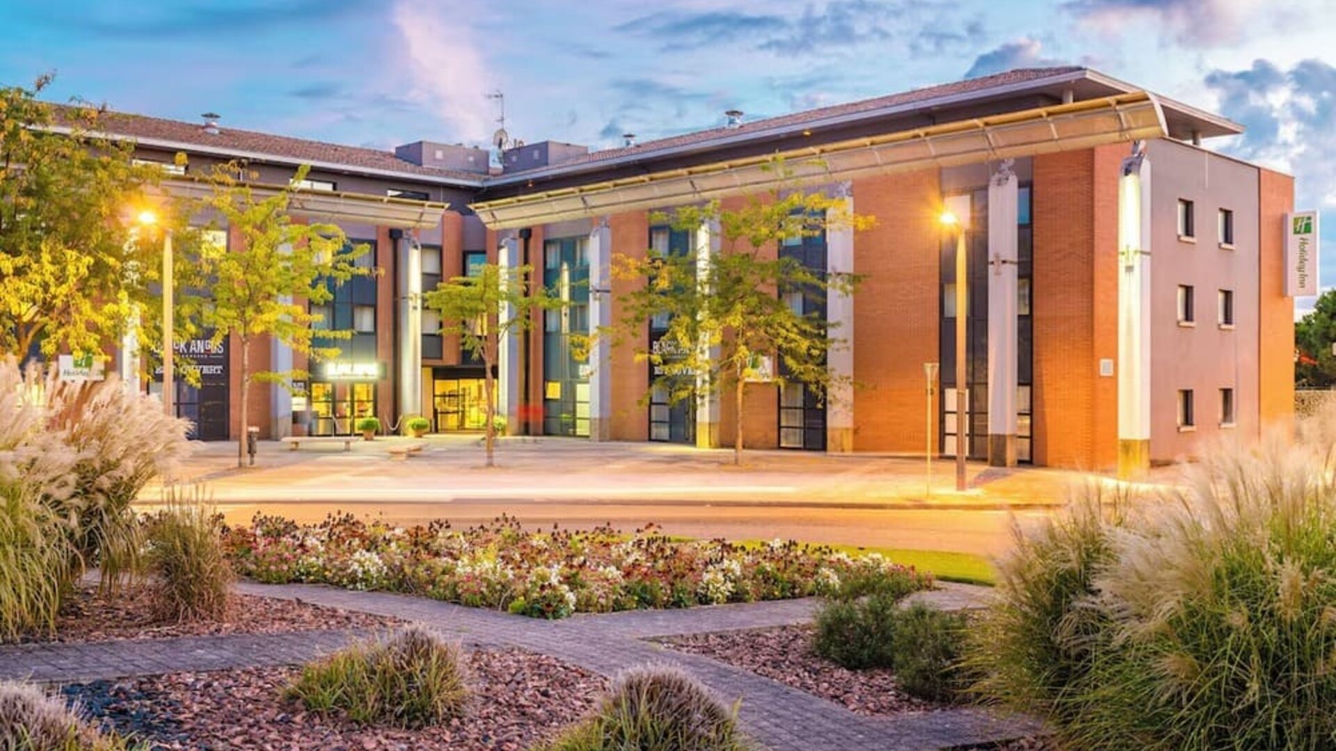 Exterior of Holiday Inn Toulouse Airport with landscaped gardens, brick and beige facade, and illuminated entrance.