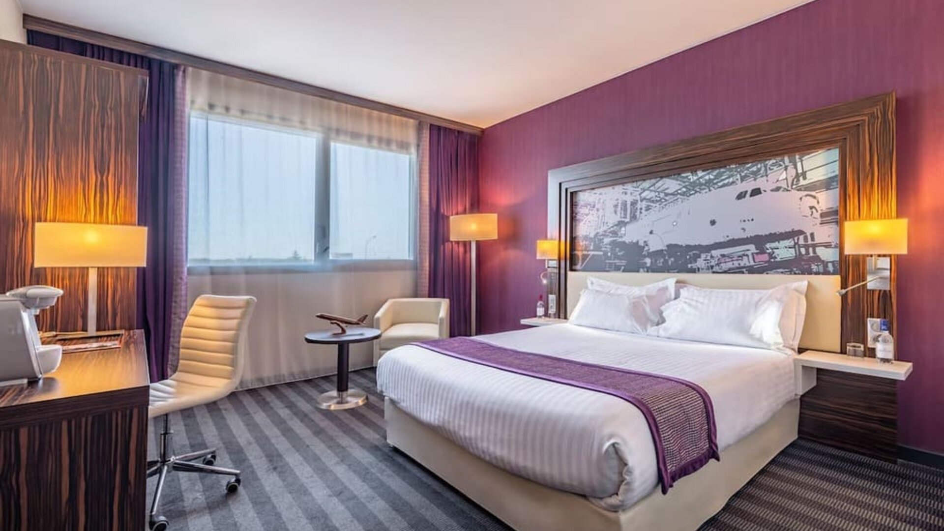 Guest room with a king bed, purple accent wall, airplane artwork, work desk, office chair, and bedside lamps at Holiday Inn Toulouse Airport.