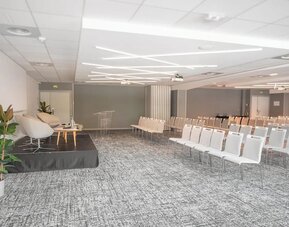 1. Large conference room with rows of white chairs, a presentation stage, podium, plants, and modern lighting at Holiday Inn Toulouse Airport.