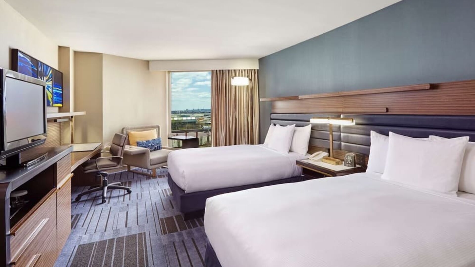 Guest room with two double beds, a blue accent wall, work desk, lounge chair, television, and large window at Hilton Newark Airport.