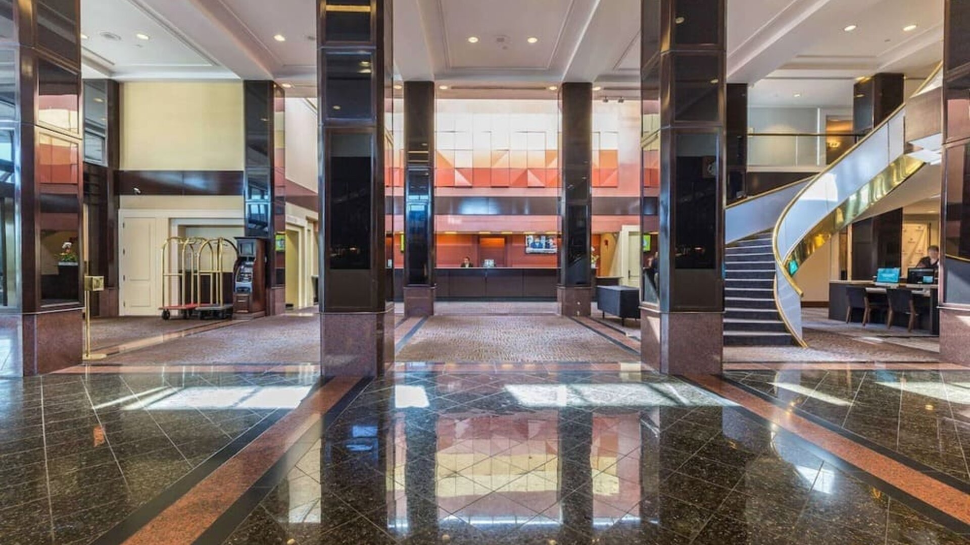 Spacious hotel lobby with polished marble floors, dark columns, reception area, luggage carts, and curved staircase at Hilton Newark Airport.