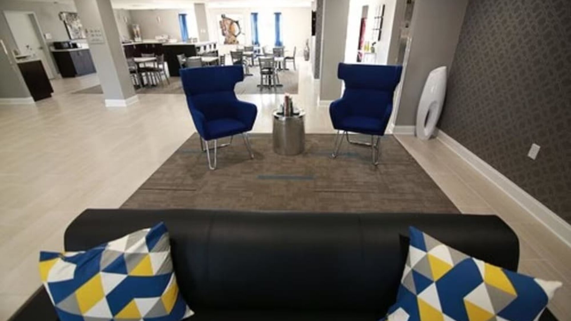 Hotel lobby seating area with a black sofa, blue armchairs, patterned accent pillows, and a dining area in the background at At Home Inn & Suites.