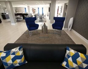 Hotel lobby seating area with a black sofa, blue armchairs, patterned accent pillows, and a dining area in the background at At Home Inn & Suites.
