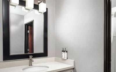 Bathroom vanity with a white countertop, oval sink, modern faucet, large framed mirror, and wall-mounted toiletries at Garner by IHG.