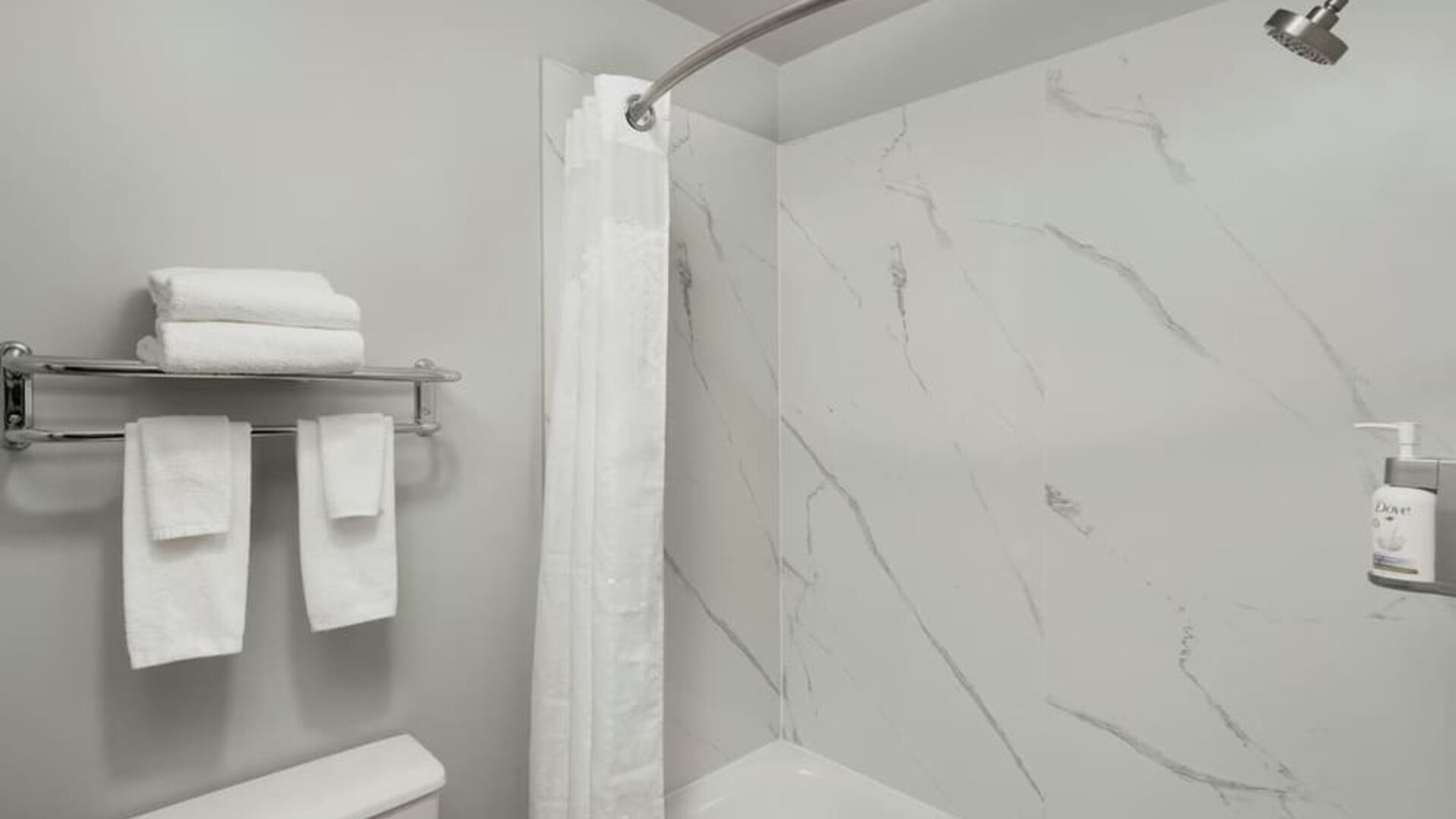 Bathroom with a bathtub and shower, white marble-look wall panels, curved shower rod, towels, and wall-mounted toiletries at Garner by IHG.