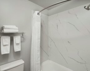 Bathroom with a bathtub and shower, white marble-look wall panels, curved shower rod, towels, and wall-mounted toiletries at Garner by IHG.