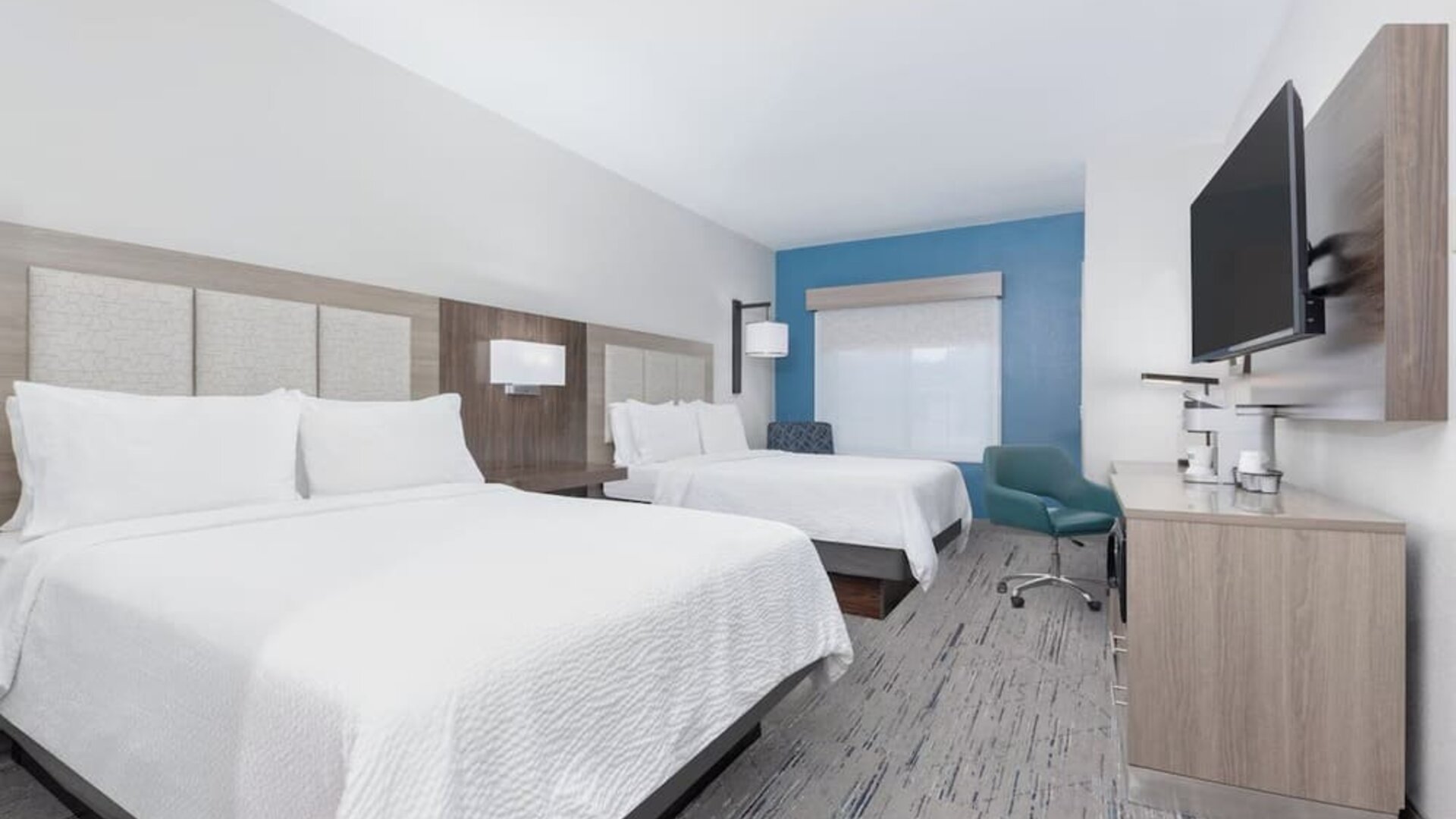 Guest room with two queen beds, white bedding, a wood-paneled headboard, blue accent wall, desk, armchair, and wall-mounted TV at Holiday Inn Express & Suites Granbury.