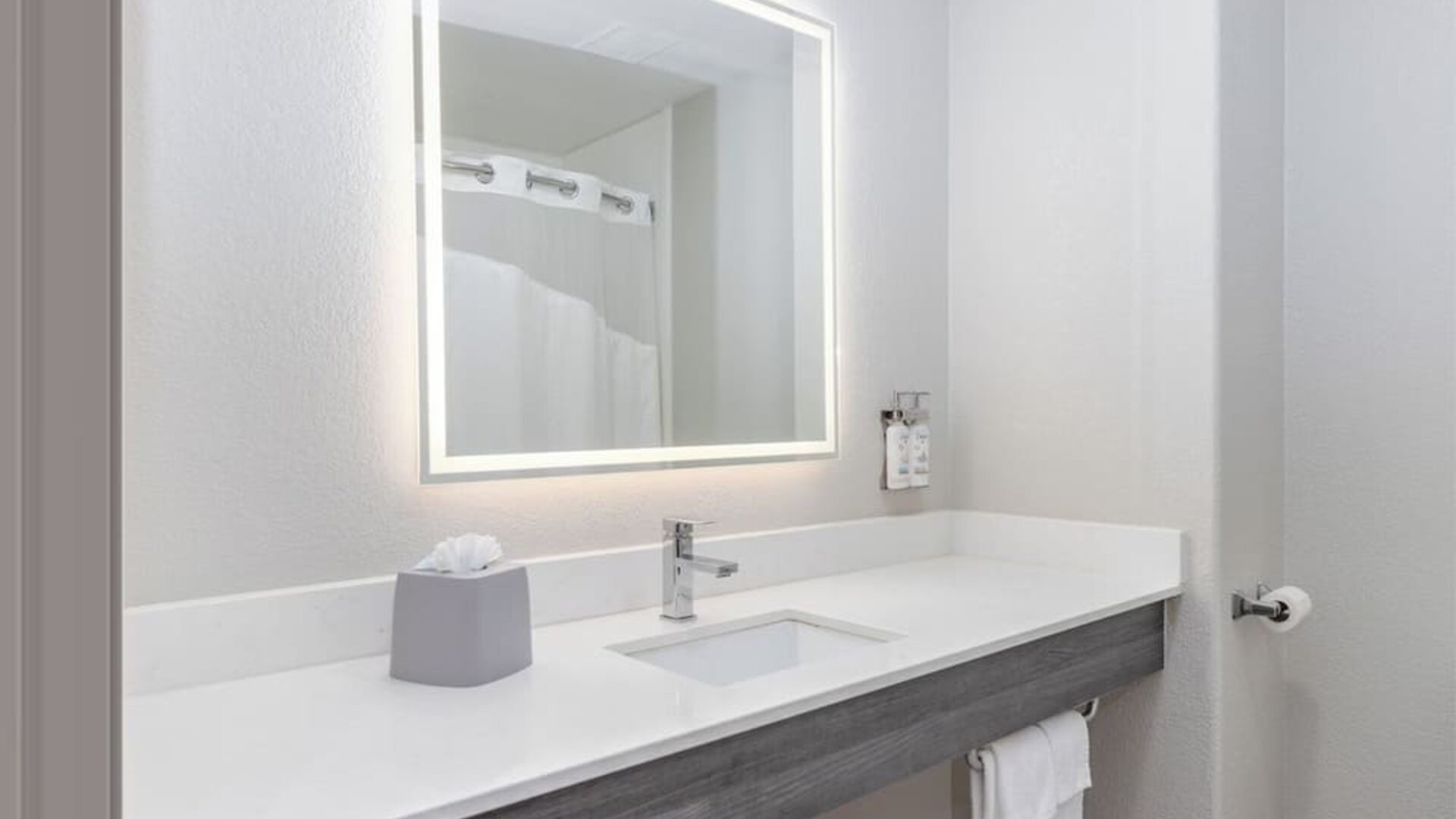 Bathroom vanity with a large illuminated mirror, white countertop, integrated sink, and wall-mounted toiletries at Holiday Inn Express & Suites Granbury.