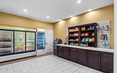 Hotel convenience and snack area with refrigerated beverages, shelves stocked with packaged snacks and travel essentials, countertop, and an ATM.