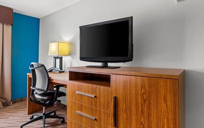 Hotel room work area with wooden desk, ergonomic office chair, large flat-screen television on a wooden cabinet, table lamp, gray wall, and blue accent wall.