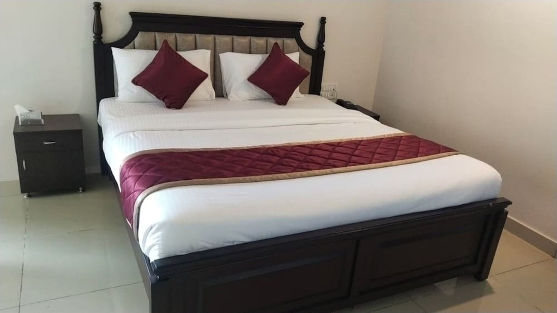 Spacious double bed featuring crisp white bedding, burgundy accent pillows, a quilted runner, an upholstered wooden headboard, and bedside furniture.