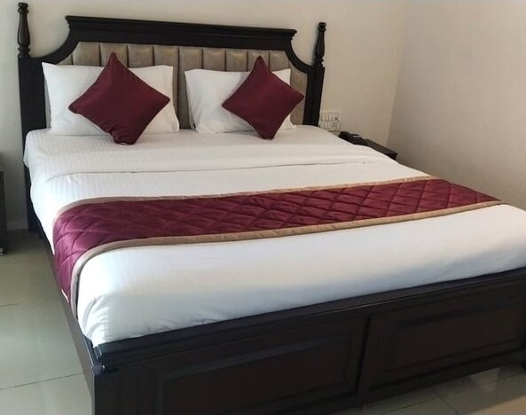 Spacious double bed featuring crisp white bedding, burgundy accent pillows, a quilted runner, an upholstered wooden headboard, and bedside furniture.
