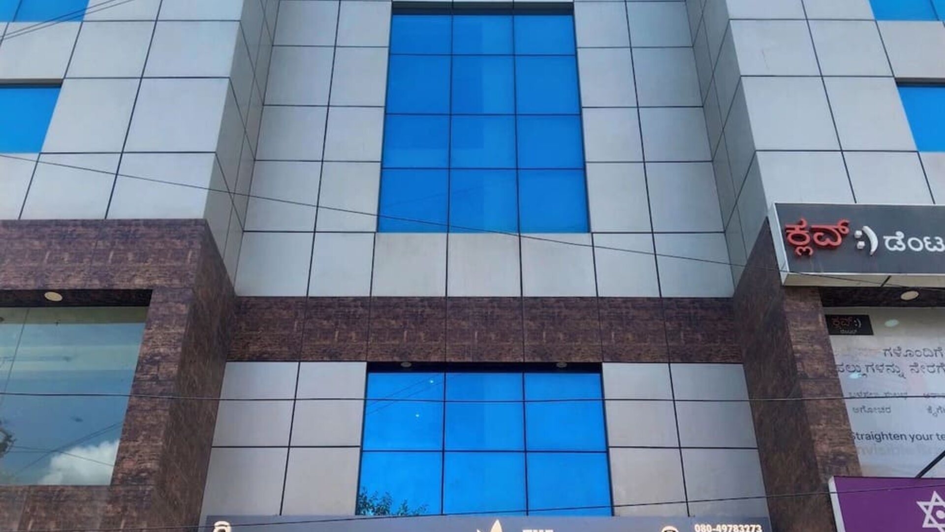 Multistory exterior of The Nisarga Suites in Bangalore featuring reflective blue glass windows, silver facade panels, brown stone accents, and hotel signage.
