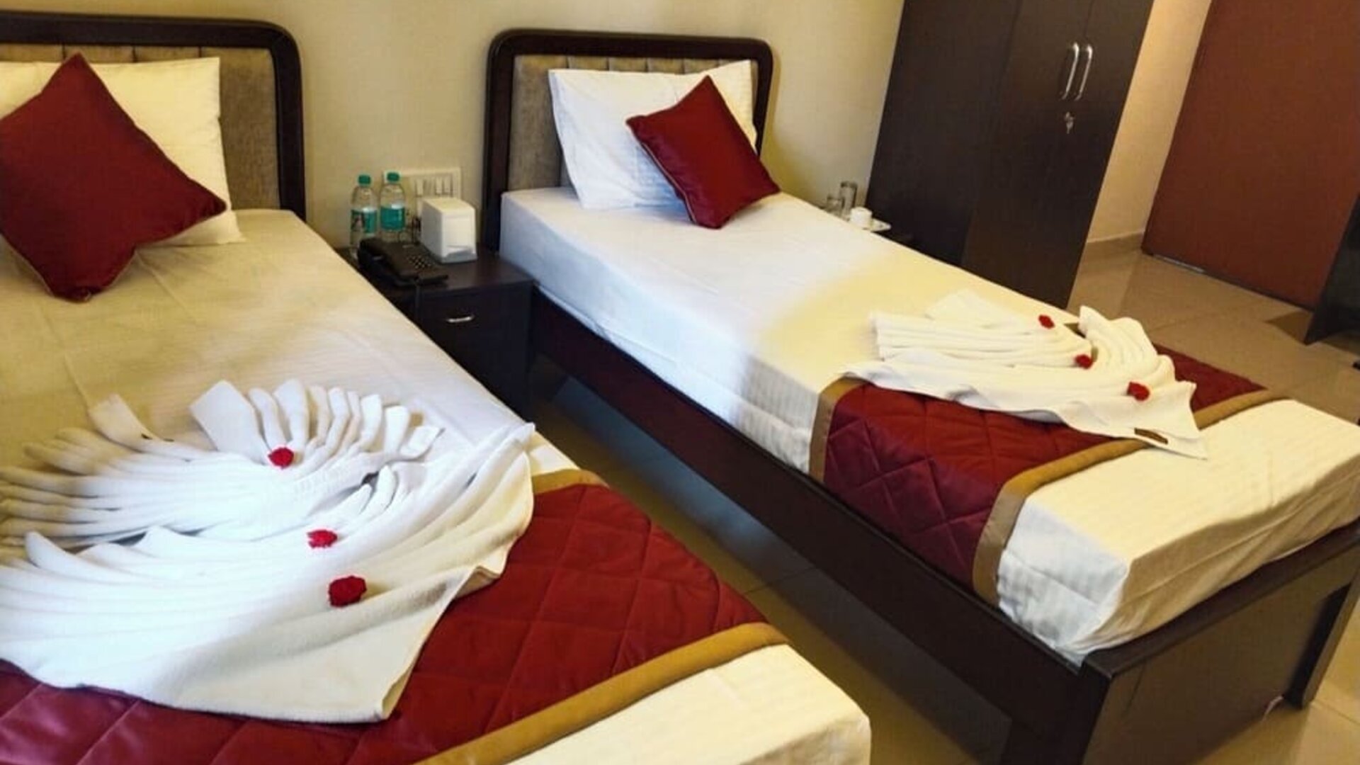 Twin bed guest room featuring white bedding, burgundy accent pillows, quilted runners, decorative folded towels with red flowers, and a bedside table with a telephone.