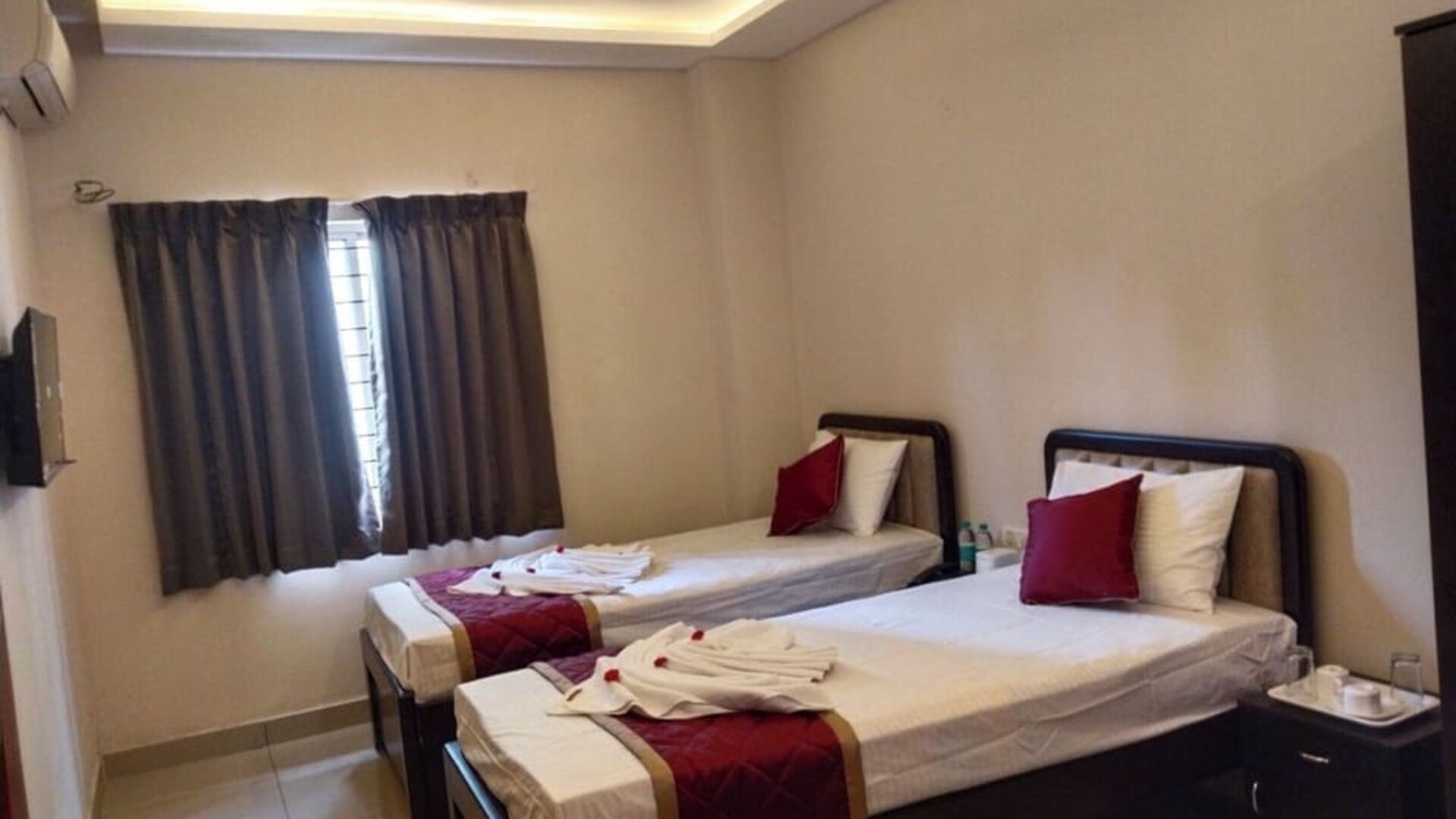 Comfortable twin bed guest room featuring white linens, burgundy decorative runners, matching pillows, folded towels, air conditioning, and a curtained window.