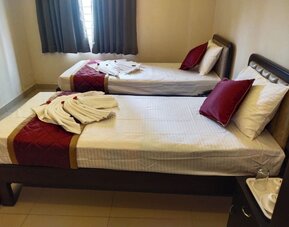 Neatly furnished twin bed guest room featuring white linens, burgundy quilted runners, decorative towel arrangements, dark wooden bed frames, and a curtained window.