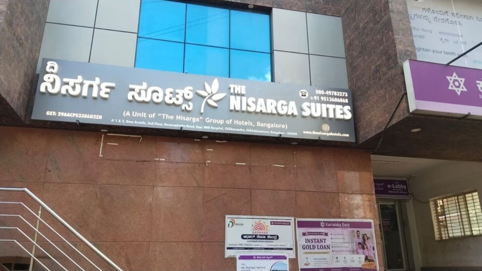Exterior entrance of The Nisarga Suites in Bangalore displaying the hotel's illuminated English and Kannada signage beneath reflective blue glass windows.