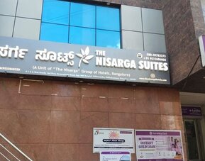 Exterior entrance of The Nisarga Suites in Bangalore displaying the hotel's illuminated English and Kannada signage beneath reflective blue glass windows.