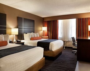 Guest room with two beds, crisp white bedding, dark bed runners, orange accent cushions, striped curtains, bedside lamps, and a work desk at Best Western Plus BWI Airport Arundel Mills.