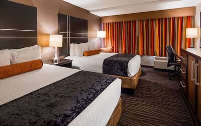 Guest room with two beds, white bedding, dark bed runners, orange accent cushions, striped curtains, bedside lamps, and a work desk at Best Western Plus BWI Airport Arundel Mills.