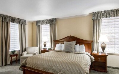 King guest room with natural window light, a traditional wooden bed, beige patterned bedding, bedside lamps, upholstered armchairs, and neutral curtains at Liberty Hotel Cleburne, an Ascend Hotel Collection Member.