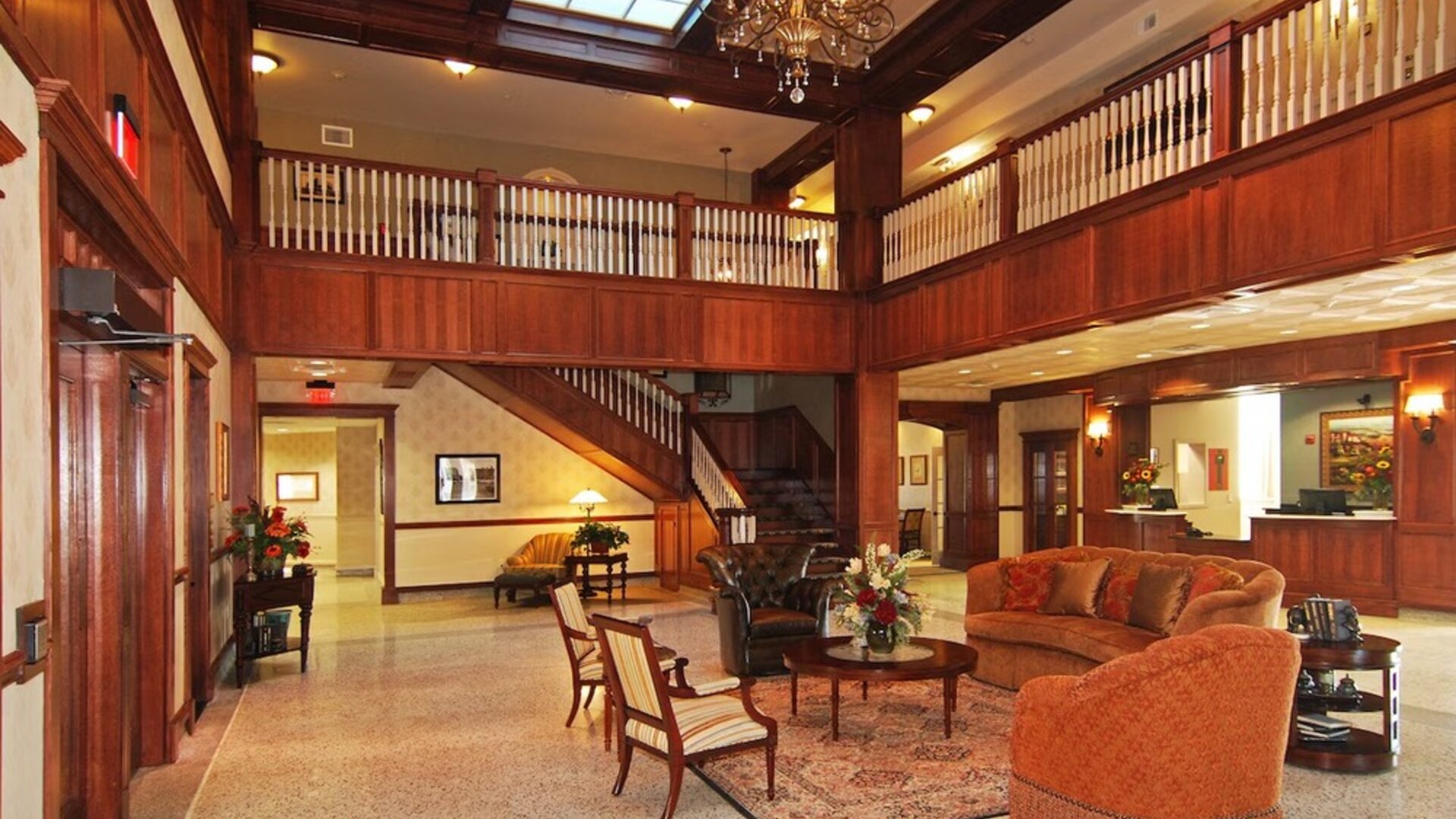 Hotel lobby with a grand wooden staircase, decorative chandelier, upper-level balcony, rich wood paneling, elegant lounge seating, and patterned rugs at Liberty Hotel Cleburne, an Ascend Hotel Collection Member.