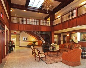 Hotel lobby with a grand wooden staircase, decorative chandelier, upper-level balcony, rich wood paneling, elegant lounge seating, and patterned rugs at Liberty Hotel Cleburne, an Ascend Hotel Collection Member.