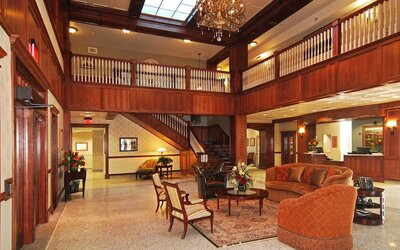Hotel lobby with a grand wooden staircase, decorative chandelier, upper-level balcony, rich wood paneling, elegant lounge seating, and patterned rugs at Liberty Hotel Cleburne, an Ascend Hotel Collection Member.