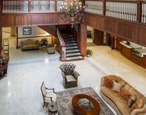 Hotel lobby lounge featuring leather armchairs, upholstered sofas, wooden furniture, a decorative chandelier, a central staircase, and a polished terrazzo floor at Liberty Hotel Cleburne, an Ascend Hotel Collection Member.